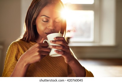 Attractive Young Black Woman Sitting At A Table At Home Backliot By A Bright Sun Enjoying A Cup Of Fresh Coffee With Her Eyes Closed In Bliss