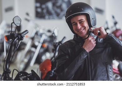 Attractive Young Blond Man In Black Leather Jacket Is Looking At Camera, Adjusting His Helmet And Smiling While Standing In A Motorbike Salon