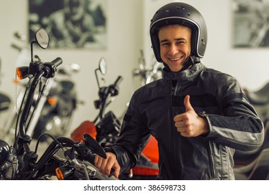 Attractive Young Blond Man In Black Leather Jacket And Helmet Is Looking At Camera, Showing Ok Sign And Smiling While Sitting On A Motorbike