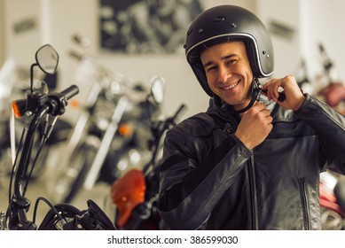 Attractive Young Blond Man In Black Leather Jacket Is Looking At Camera, Adjusting His Helmet And Smiling While Standing In A Motorbike Salon