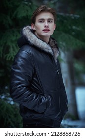 Attractive young blond man in stylish black leather jacket with a fur hood stands in a winter park covered with snow holding his hands in pockets. Men's winter fashion.