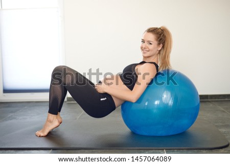 Attractive young blond pregnant woman working out with a pilates ball in a gym holding her swollen belly with a smile as she balances on her toes to strengthen her abdominal muscles
