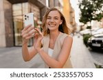 attractive young blond woman sitting in city street using smart phone in spring summer autumn fashion trend outfit smiling happy wearing beige suit and handbag style in sunny warm tones