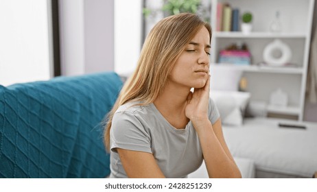 Attractive young blonde adult woman sitting on living room sofa at home, suffering in agony from severe teeth pain - oral issue demanding dentist's help urgently. - Powered by Shutterstock