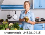 Attractive young blonde woman holding jar of pickles looking at camera, cooking salad in modern kitchen, preparing vegetarian meal, keeps diet, wearing blue dress.