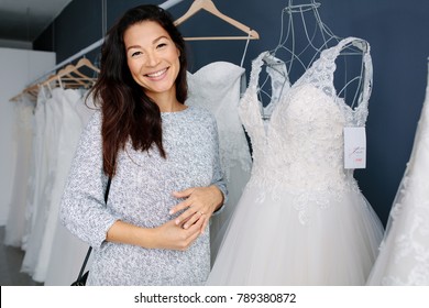 Attractive Young Bride Smiling While Choosing Wedding Dress In Bridal Boutique. Smiling Asian Woman Shopping For Wedding Outfit In Bridal Clothing Store.