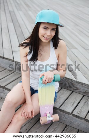 Attractive young brunette girl sitting outdoors with short cruiser penny skate board at summer day. Popular small skate deck, trendy outfit, beautiful model