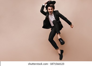 Attractive young brunette having fun and posing extraordinary against beige background. Man wearing white shirt, black striped suit, classic hat and elegant shoes