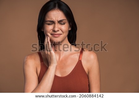 Attractive young brunette woman with bad tooth ache holding her hand to her jaw with her mouth open as she frowns in pain, isolated on beige color background