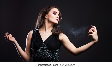 Attractive Young Brunette Woman And A Bottle With A New Fragrance, Use Perfume. Portrait On A Black Background