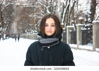 An attractive young brunette woman stands in snow-covered winter park smiles looking at a camera. Portrait of a beautiful female girl. A path covered with white snow goes into distance. Cold weather