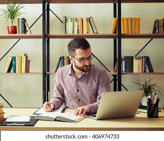 Attractive young businessman with the glasses rewriting from a book and looking at his laptop