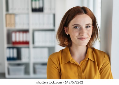 Attractive young businesswoman with a happy smile leaning against an office wall looking at the camera in a close up portrait