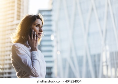 Attractive young businesswoman wearing white shirt and talking on smartphone with business partners outdoors, professional female architect working on new project of office buildings, flare light