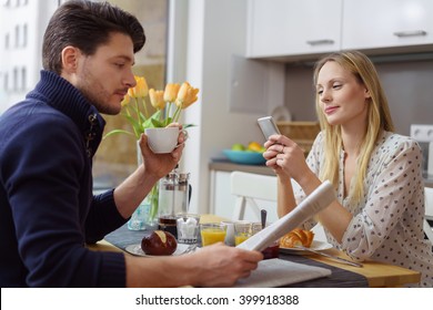 Attractive Young Caucasian Couple Distracted At Table With Newspaper And Cell Phone While Eating Breakfast