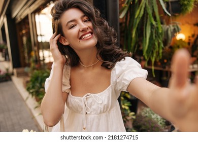 Attractive Young Caucasian Girl With Toothy Smile Looks At Camera Standing On Street. Brunette Straightens Her Hair Behind Ear, Wears Sundress In Summer. Sincere Emotions Lifestyle Concept