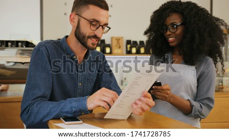 Attractive young caucasian man making an order in the cafe in the african woman using menu, she writing down everything in notebook.