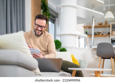 Attractive Young Caucasian Manager Working From Home Comfortably Sitting In His Sofa Taking Note And Comparing Business Results. Concept Of New Way Of Living