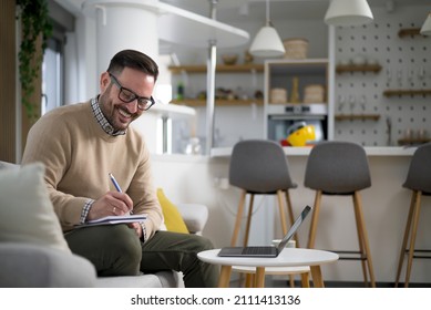 Attractive Young Caucasian Manager Working From Home Comfortably Sitting In His Sofa Taking Note And Comparing Business Results. Concept Of New Way Of Living