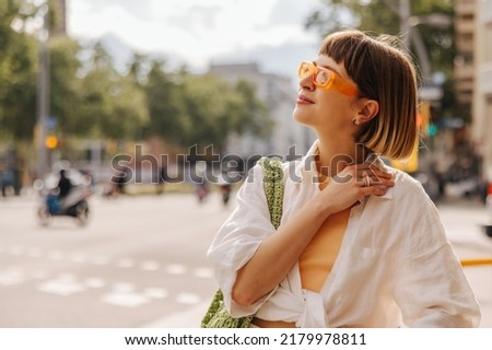 Attractive young caucasian woman is enjoying warm sunny weather walking outside. Brown-haired with bob haircut wears sunglasses, shirt. Relaxation concept