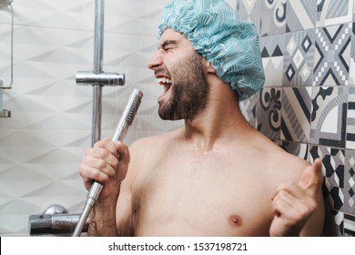 Attractive young cheerful man singing while washing in the shower, wearing shower cap and holding shower head - Powered by Shutterstock