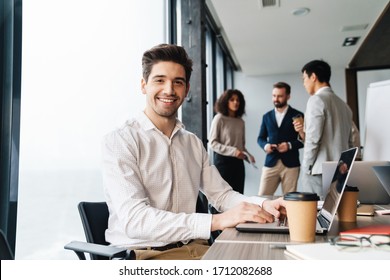 Attractive Young Confident Businessman Sitting At The Office Table With Group Of Colleagues In The Background, Working On Laptop Computer