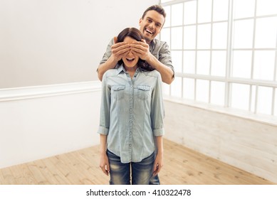 Attractive Young Couple Is Moving, Standing In An Empty New Apartment. Man Is Covering His Woman Eyes Making A Surprise