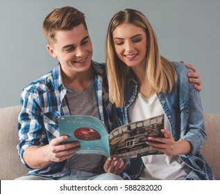 Attractive Young Couple Is Reading A Magazine, Hugging And Smiling While Sitting On Couch At Home