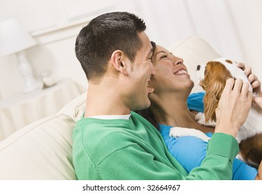 An Attractive Young Couple Sitting On A Couch Together And Holding A Dog.  They Are Laughing.  Horizontally Framed Shot.