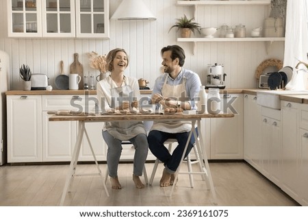Attractive young couple wear aprons spend time together in the kitchen, knead dough for home-made pastries, buns or dumplings, prepare biscuits for family holiday. Family recipe and traditions, date