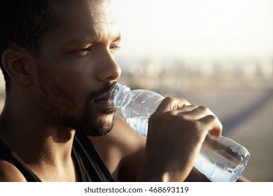 Attractive young dark skinned sportsman with short beard drinking water from bottle looking far away with thoughtful face expression, dressed in black sleeveless shirt, relaxing after morning run