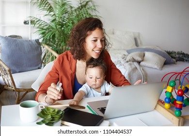 Attractive young dark skinned woman working at desk at home using laptop, holding baby on her lap. Portrait of smiling mother writing post on moms blog while her infant son playing with toy