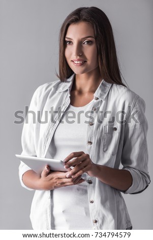 Attractive young dark-haired woman in casual clothes is using a digital tablet and smiling, on gray background