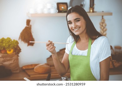 An attractive young dark-haired woman tasting ready hot meal with a wooden spoon while standing and smiling in sunny kitchen. Cooking and householding concepts - Powered by Shutterstock