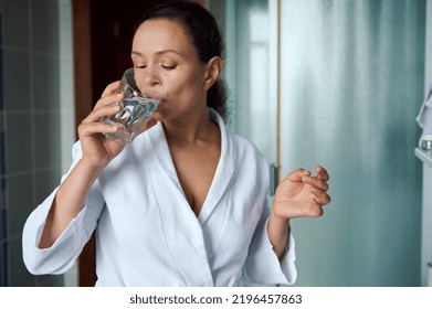 Attractive Young Dark-haired Woman In White Bathrobe, Holding Pill And Glass Of Fresh Pure Water, Taking Antioxidant Medicine Vitamins, Beauty Supplements For Hair Skin Nails. Healthcare Concept.