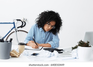 Attractive young dark-skinned female columnist suffering from writer's block, working on new article for online women's magazine, making notes in sketchbook, surrounded by crumpled paper balls - Powered by Shutterstock