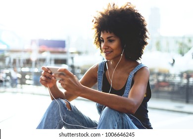 An Attractive Young Dark-skinned Girl Looking At Video On Mobile Phone While Relaxing Outside In Summer Warm Day. Young Girl With Afro Hair Playing In A Game On The Cellphone. Girl  Watching Videos