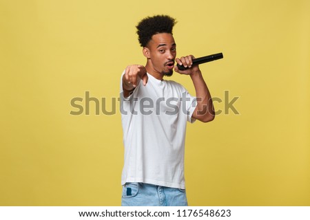 Attractive young dark-skinned man with afro haircut in white t shirt, gesticulating with hands and microphone, dancing and singing on party, having fun.