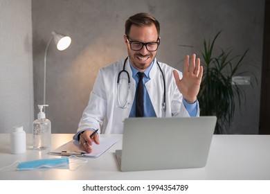 Attractive Young Doctor Having Online Appointment With His Patient. He Is Looking At Laptop, Smiling And Saying Hi