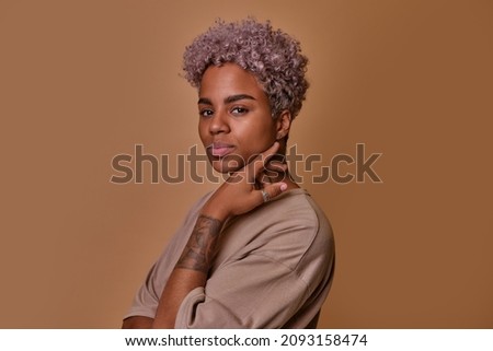 Attractive young ethnic African American woman confidently looking at camera and smiling slightly, stylish girl with tattoo on arm posing in studio on brown background. Lifestyle people portrait