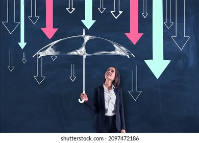 Attractive young european business woman holding drawn umbrella sketch to protect herself from colorful downward arrows on chalkboard wall background. Finance, investment and insurance concept - Powered by Shutterstock