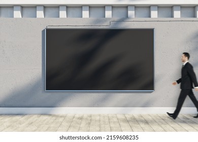 Attractive Young European Businessman Walking Past Empty Black Poster Hanging On Concrete Outdoor Wall With Shadows And Sunlight. Billboard, Commercial And Advertisement Concept