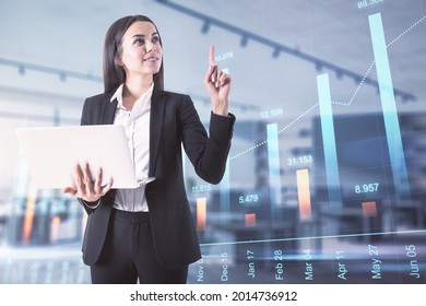 Attractive young european businesswoman with laptop and abstract forex chart standing on blurry background with mockup place. Technology, trade and finance concept. Double exposure