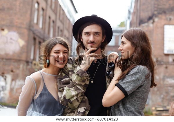 Attractive young European man wearing trendy hat\
and camouflage jacket relaxing in urban setting with his two\
girlfriends, enjoying good company. Three cheerful teenage friends\
having fun outdoor