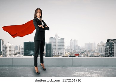 Attractive young european woman with red hero cape and folded arms standing on city background. Superhero and success concept 
