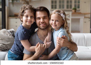 Attractive Young Father And Little Kids Looking At Camera Seated On Couch In Living Room, Siblings Embracing Loving Step Dad Showing Love And Sincere Feelings, Concept Of Single Parent Or Sole Custody