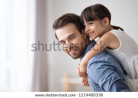 Attractive young father standing in living room holds on back adorable daughter at home. Smiling daughter spend time with loving father playing piggyback her daddy. Family leisure activities concept