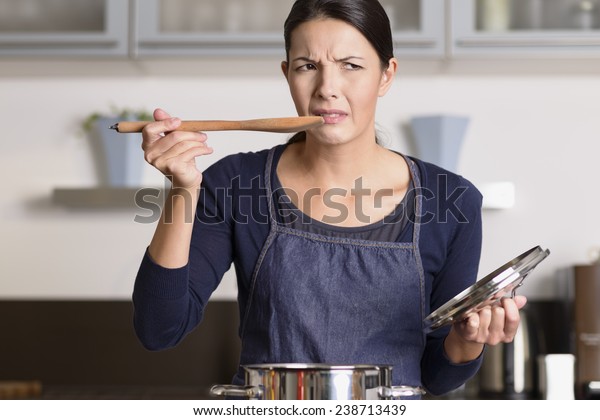Attractive young female cook standing at the\
hob in her apron tasting her food in the saucepan with a grimace as\
she finds it distasteful and\
unpalatable