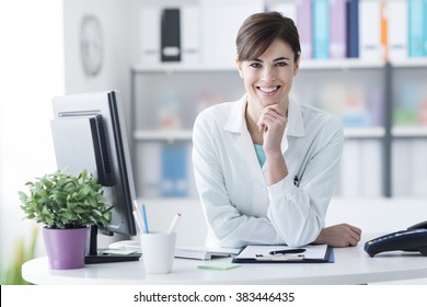 Attractive Young Female Doctor Leaning On The Clinic Reception Desk With Hand On Chin, She Is Smiling At Camera, Medical Staff And Healthcare Concept