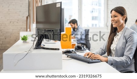 Attractive young female employee worker in suit using computer while sitting at her desk in office.